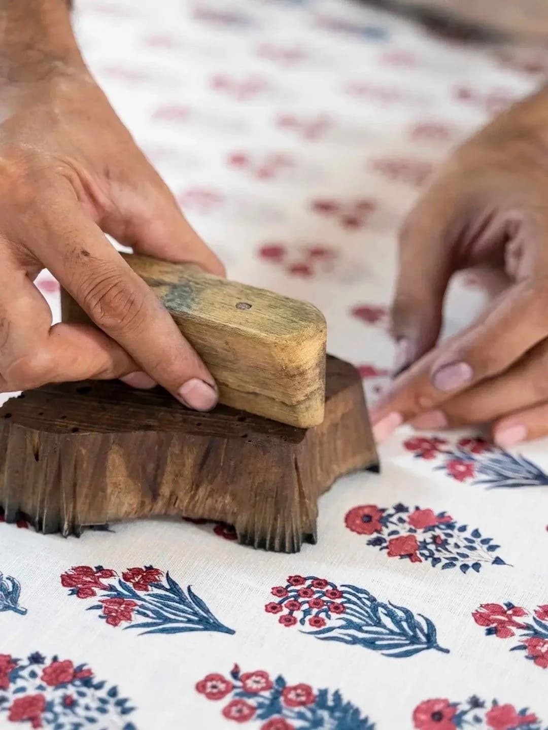An artisan's hands pressing a carved wooden block onto cotton, printing rows of red and indigo florals.
