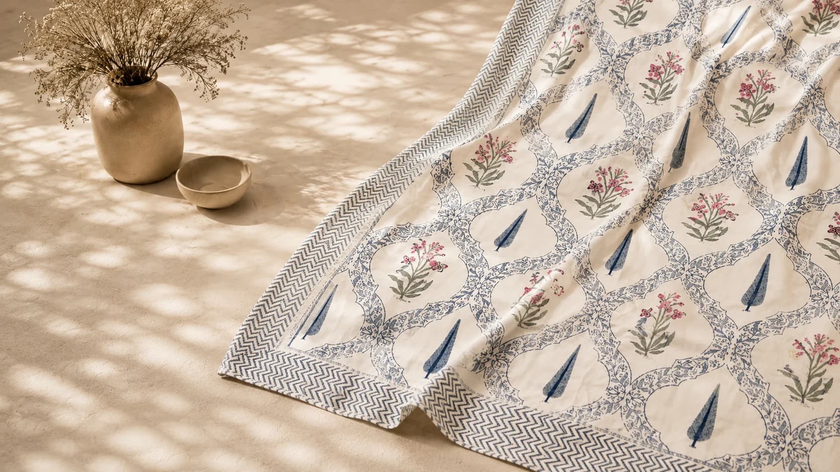 Hand block-printed cotton in indigo and rose florals, laid out beside a ceramic vase of dried blooms.