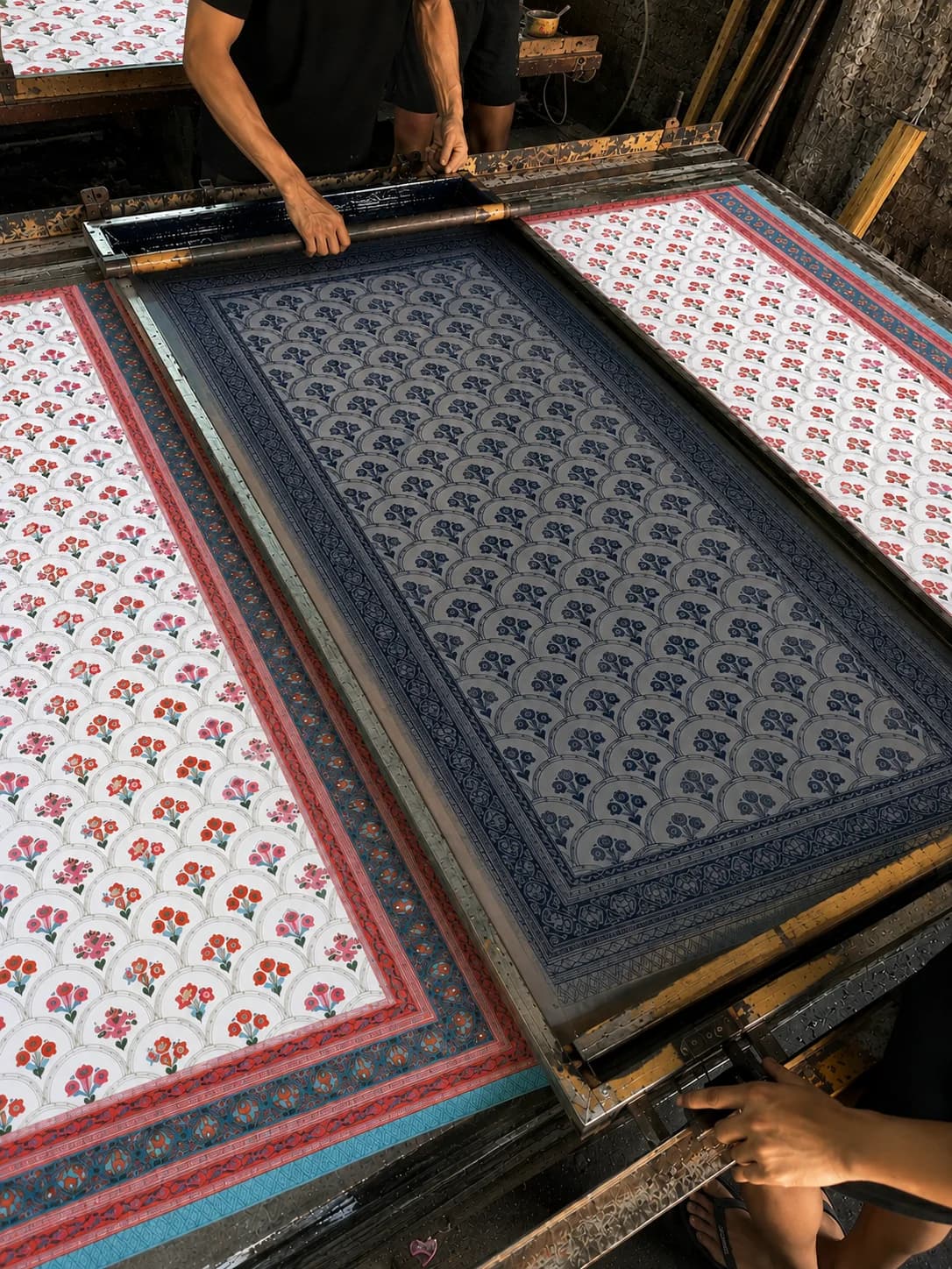 A printer pulling ink across a screen over cotton in a Jaipur workshop, panels of finished floral prints laid out alongside.