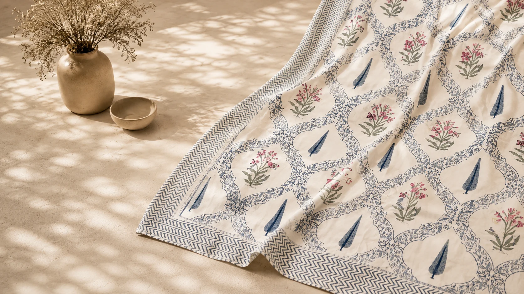 Hand block-printed cotton in indigo and rose florals, laid out beside a ceramic vase of dried blooms.