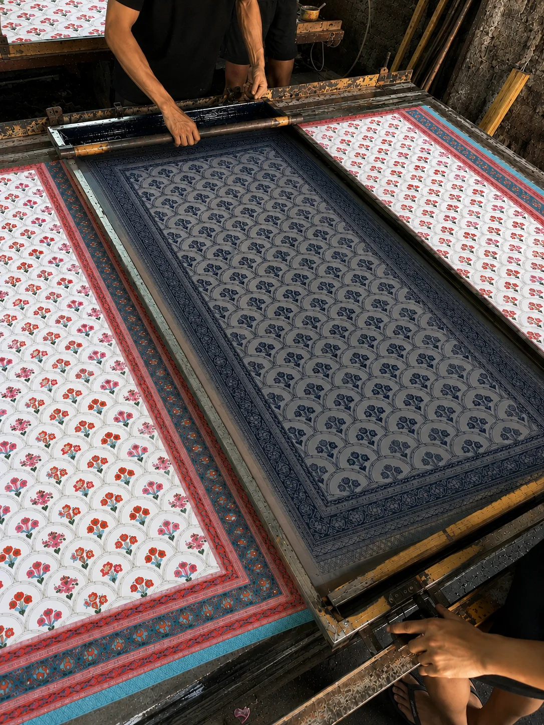 A printer pulling ink across a screen over cotton in a Jaipur workshop, panels of finished floral prints laid out alongside.